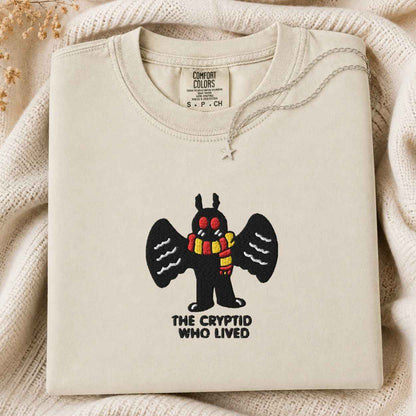 A folded cream-colored tshirt with a graphic of a black cartoon mothman cryptid with red eyes and a yellow-and-red striped scarf, above the text 'THE CRYPTID WHO LIVED'. The tshirt is styled on a textured cream knit blanket with dried flowers in the corner.