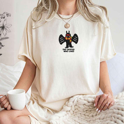 A woman wearing a cream-colored oversized t-shirt with a graphic of a winged cryptid and the text 'THE CRYPTID WHO LIVED'. She is holding a white mug and sitting on a white bed with a chunky knit blanket.