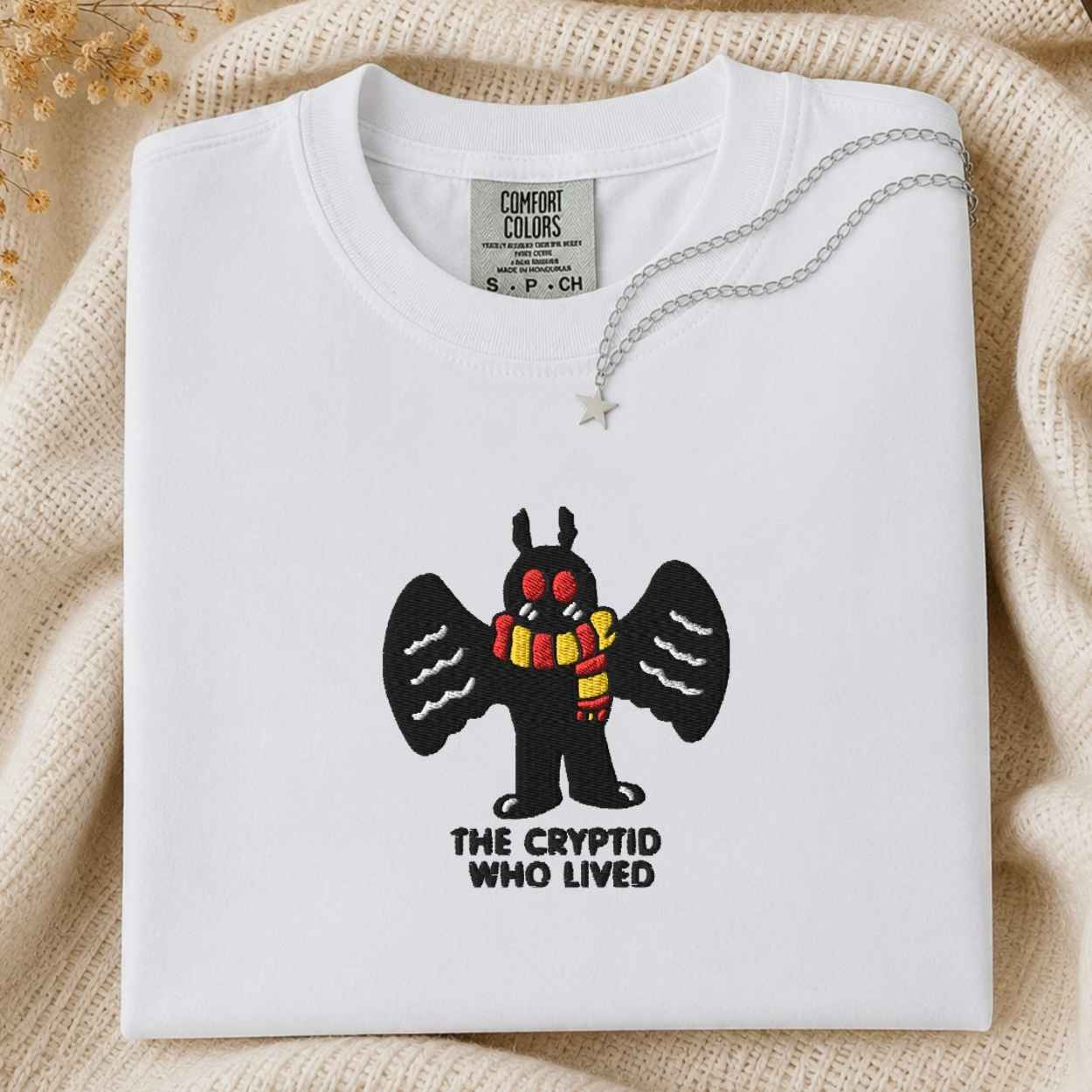 A white Comfort Colors t-shirt featuring a cartoon cryptid with black wings and a red and yellow striped scarf, with the text 'THE CRYPTID WHO LIVED' underneath. The shirt is styled flat on a beige knit blanket with a silver necklace draped over the collar.