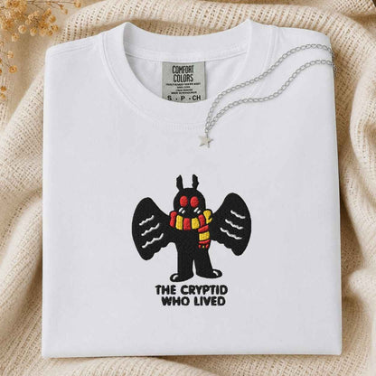 A white Comfort Colors t-shirt featuring a cartoon cryptid with black wings and a red and yellow striped scarf, with the text 'THE CRYPTID WHO LIVED' underneath. The shirt is styled flat on a beige knit blanket with a silver necklace draped over the collar.