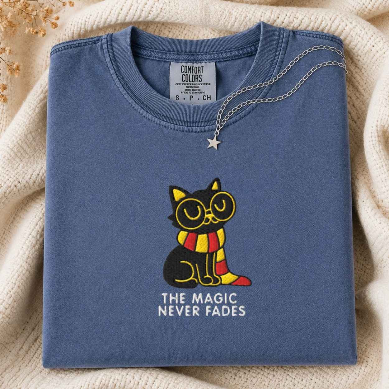 A folded slate blue t-shirt with a graphic of a black cat wearing round glasses and a red and yellow striped scarf, with the text 'THE MAGIC NEVER FADES' underneath. A silver necklace with a star is draped over the collar.
