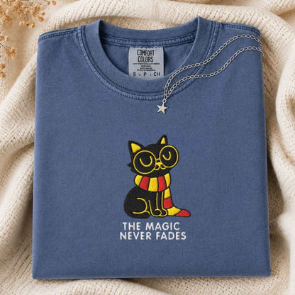 A folded slate blue t-shirt with a graphic of a black cat wearing round glasses and a red and yellow striped scarf, with the text 'THE MAGIC NEVER FADES' underneath. A silver necklace with a star is draped over the collar.