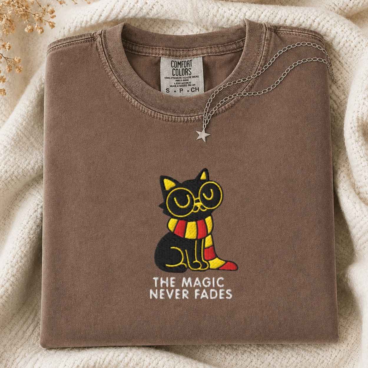 A folded brown t-shirt with a graphic of a black cat wearing round glasses and a red and yellow striped scarf, with the text 'THE MAGIC NEVER FADES' underneath. The t-shirt is styled on a cream-colored knit blanket with a silver star pendant necklace.