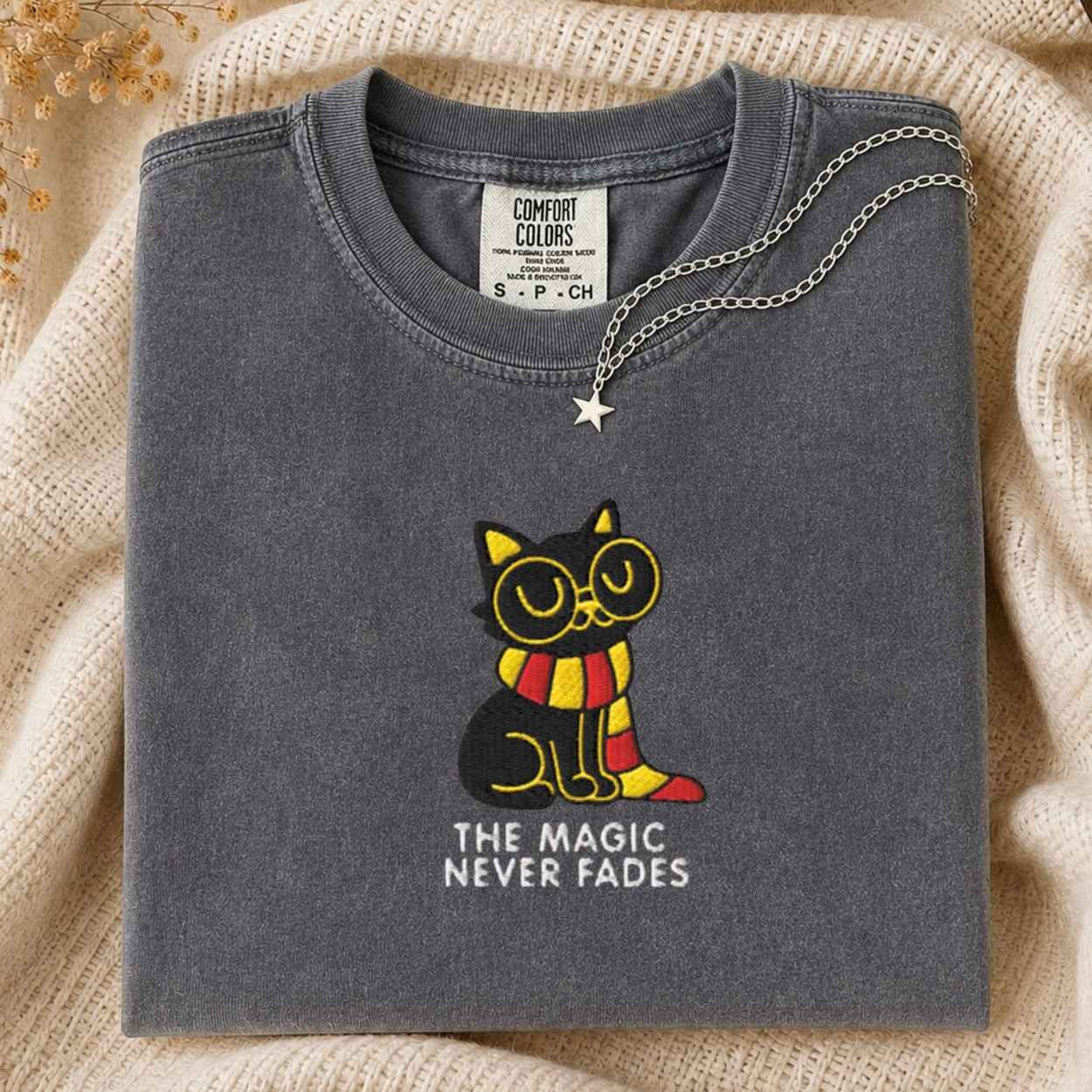 A folded dark gray t-shirt with a graphic of a black cat wearing yellow glasses and a red and yellow striped scarf, with the text 'THE MAGIC NEVER FADES' underneath. A silver star necklace is draped over the collar.