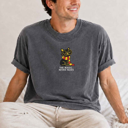 A man wearing a dark gray washed t-shirt with a graphic of a cat in a yellow and red striped scarf and the text 'THE MAGIC NEVER FADES'.