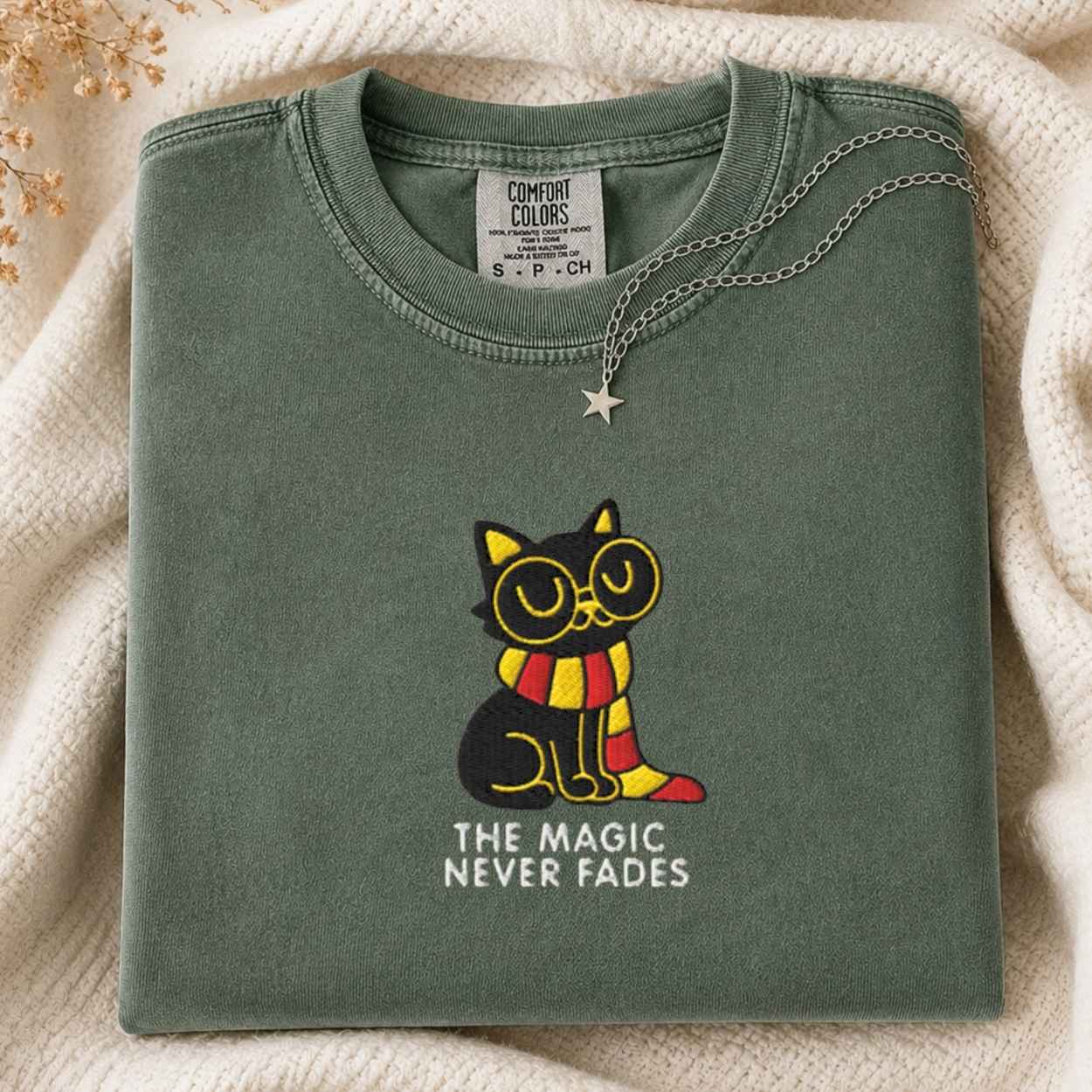 A folded sage green t-shirt with a graphic of a black cat wearing round glasses and a red and yellow striped scarf, with the text 'THE MAGIC NEVER FADES' underneath. The t-shirt is styled on a cream knit blanket with a silver star necklace draped over it.
