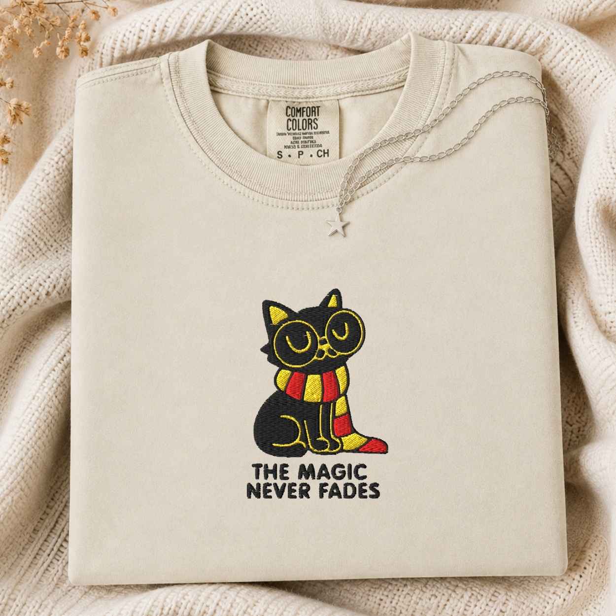 A folded cream-colored tshirt from the brand 'Comfort Colors' is displayed on a cozy, textured beige knit blanket. The tshirt features a graphic of a black cat wearing yellow glasses and a red-and-yellow striped scarf, with the text 'THE MAGIC NEVER FADES' printed below.