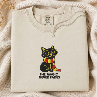 A folded cream-colored tshirt from the brand 'Comfort Colors' is displayed on a cozy, textured beige knit blanket. The tshirt features a graphic of a black cat wearing yellow glasses and a red-and-yellow striped scarf, with the text 'THE MAGIC NEVER FADES' printed below.