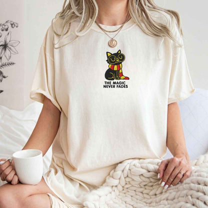 A woman wearing a cream-colored oversized t-shirt with a graphic of a black cat wearing a red and yellow striped scarf and the text 'THE MAGIC NEVER Fades'. She is sitting on a bed with a chunky knit blanket and holding a white mug.