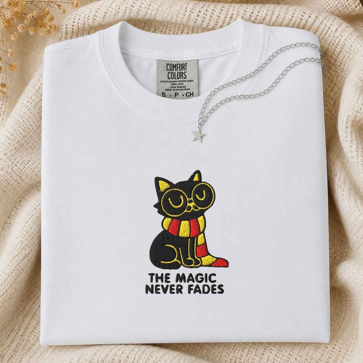 A folded white Comfort Colors t-shirt with a graphic of a black cat wearing yellow glasses and a red and yellow striped scarf, with the text 'THE MAGIC NEVER FADES' underneath. The shirt is styled on a beige knit blanket with a silver necklace draped over the collar.