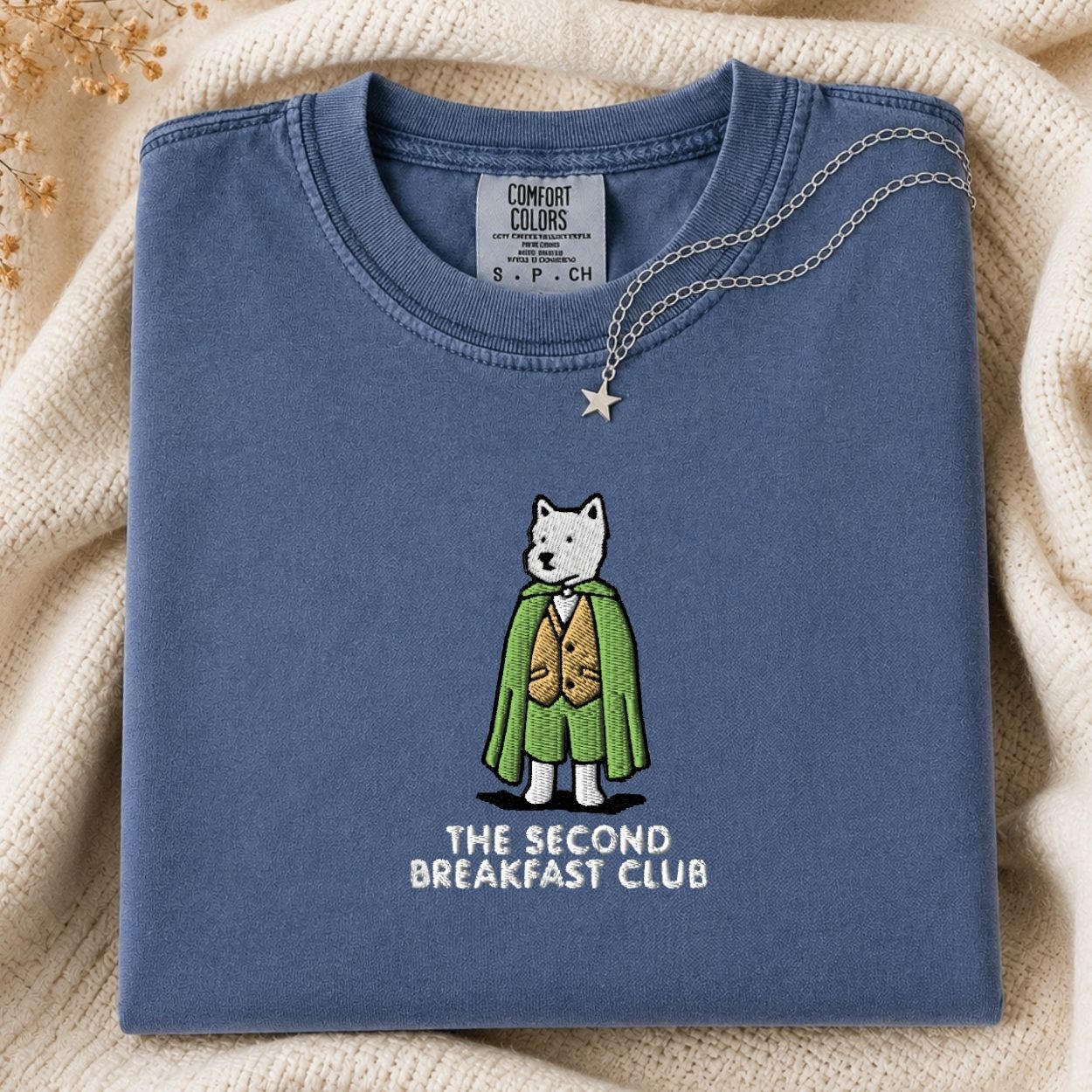A folded slate blue t-shirt with a graphic of a dog character in a green cape and gold vest, with the text 'THE SECOND BREAKFAST CLUB' printed below. The shirt is styled on a cream-colored knit blanket with a gold necklace draped over the collar.