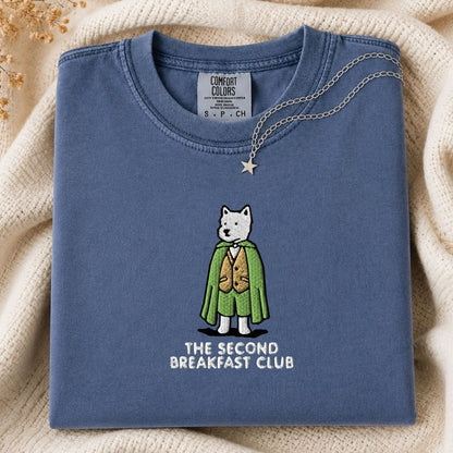 A folded slate blue t-shirt with a graphic of a dog character in a green cape and gold vest, with the text 'THE SECOND BREAKFAST CLUB' printed below. The shirt is styled on a cream-colored knit blanket with a gold necklace draped over the collar.