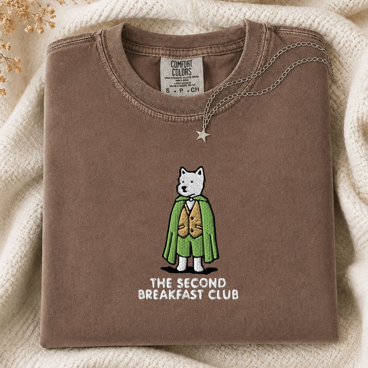 A folded brown tshirt with a graphic of a white dog in a green and gold outfit and the text 'THE SECOND BREAKFAST CLUB' on the front, styled on a cream knit blanket with a silver necklace.
