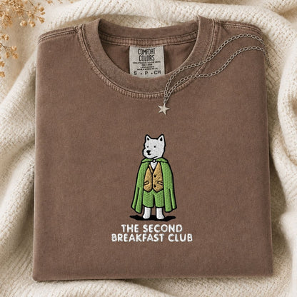 A folded brown tshirt with a graphic of a white dog in a green and gold outfit and the text 'THE SECOND BREAKFAST CLUB' on the front, styled on a cream knit blanket with a silver necklace.