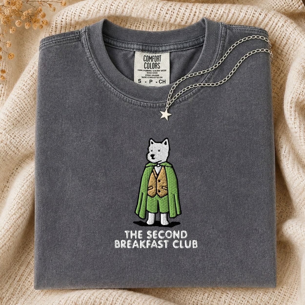 A folded dark gray t-shirt with a graphic of a white dog in a green cape and brown vest, with the text 'THE SECOND BREAKFAST CLUB' underneath. The shirt is styled on a beige knit blanket with a silver necklace draped over the collar.