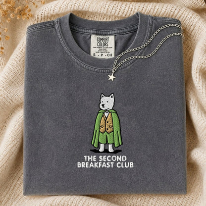 A folded dark gray t-shirt with a graphic of a white dog in a green cape and brown vest, with the text 'THE SECOND BREAKFAST CLUB' underneath. The shirt is styled on a beige knit blanket with a silver necklace draped over the collar.