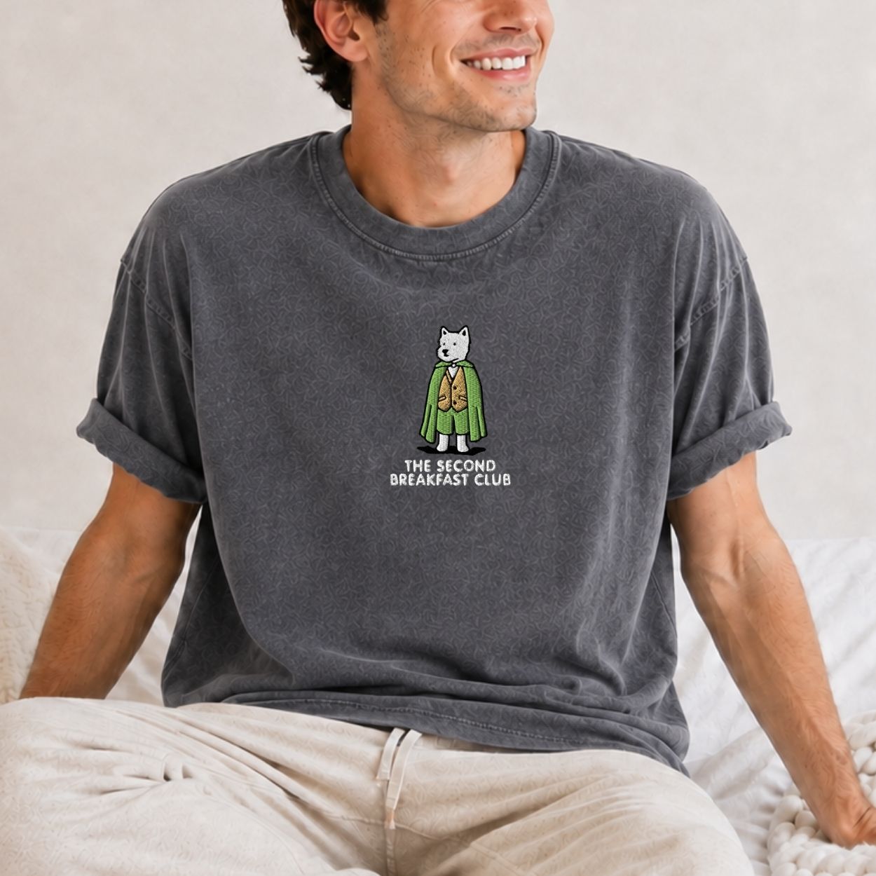 A man wearing a dark gray heathered t-shirt with a cartoon dog character wearing a robe and the text 'the second breakfast club' printed on the chest, sitting on a bed.