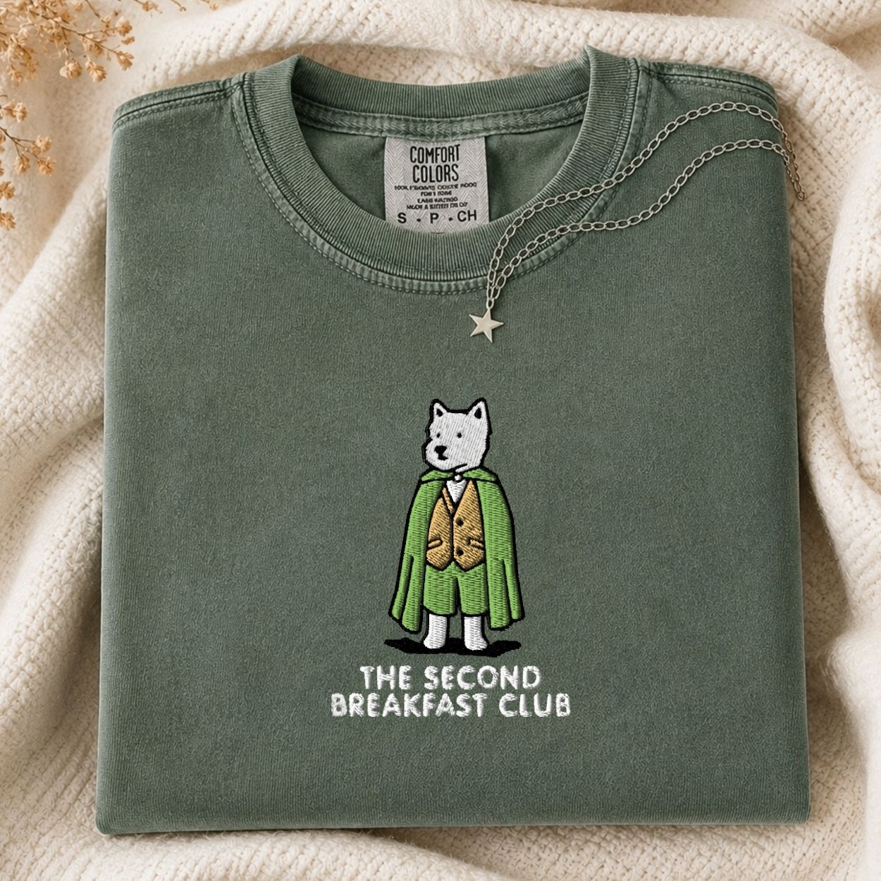 A folded sage green t-shirt with a graphic of a white dog in a green cape and gold vest, with the text 'THE SECOND BREAKFAST CLUB' underneath. The shirt is styled on a cream-colored knit blanket with a star necklace draped over the collar.