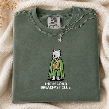 A folded sage green t-shirt with a graphic of a white dog in a green cape and gold vest, with the text 'THE SECOND BREAKFAST CLUB' underneath. The shirt is styled on a cream-colored knit blanket with a star necklace draped over the collar.