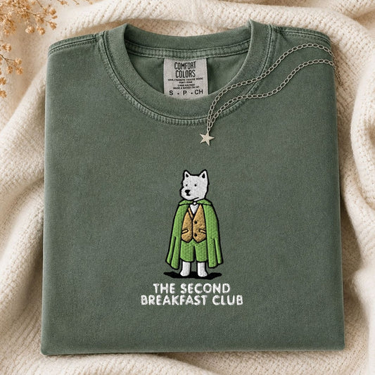 A folded sage green t-shirt with a graphic of a white dog in a green cape and gold vest, with the text 'THE SECOND BREAKFAST CLUB' underneath. The shirt is styled on a cream-colored knit blanket with a star necklace draped over the collar.