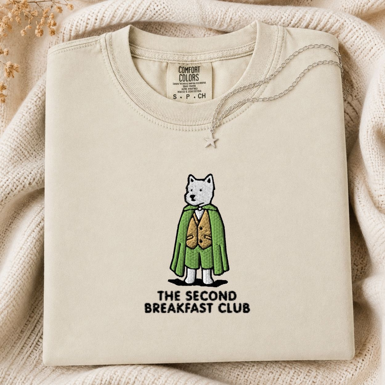 A folded cream-colored crewneck tshirt with a graphic of a white dog in a green cape and brown vest, with the text 'THE SECOND BREAKFAST CLUB' underneath. The tshirt is displayed on a cozy beige knit blanket with dried flowers in the corner.