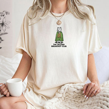 A woman wearing an oversized cream-colored t-shirt with a graphic of a dog in a green cloak and the text 'THE SECOND BREAKFAST CLUB'. She is holding a white mug and sitting on a cozy knit blanket.