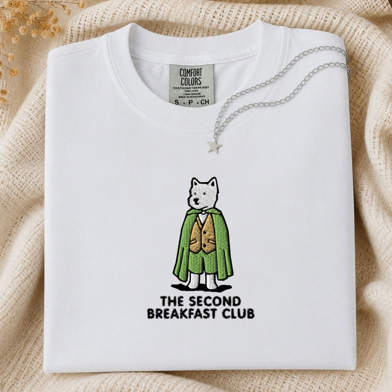 A folded white Comfort Colors t-shirt featuring a graphic of a dog dressed in a green cape and gold vest with the text 'THE SECOND BREAKFAST CLUB' underneath. The shirt is styled on a beige knit blanket with a silver necklace draped over the collar.