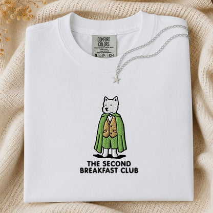 A folded white Comfort Colors t-shirt featuring a graphic of a dog dressed in a green cape and gold vest with the text 'THE SECOND BREAKFAST CLUB' underneath. The shirt is styled on a beige knit blanket with a silver necklace draped over the collar.